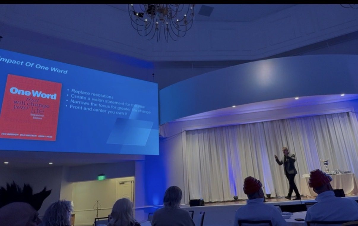 Speaker presenting on stage with projected slide to an audience in a conference room.