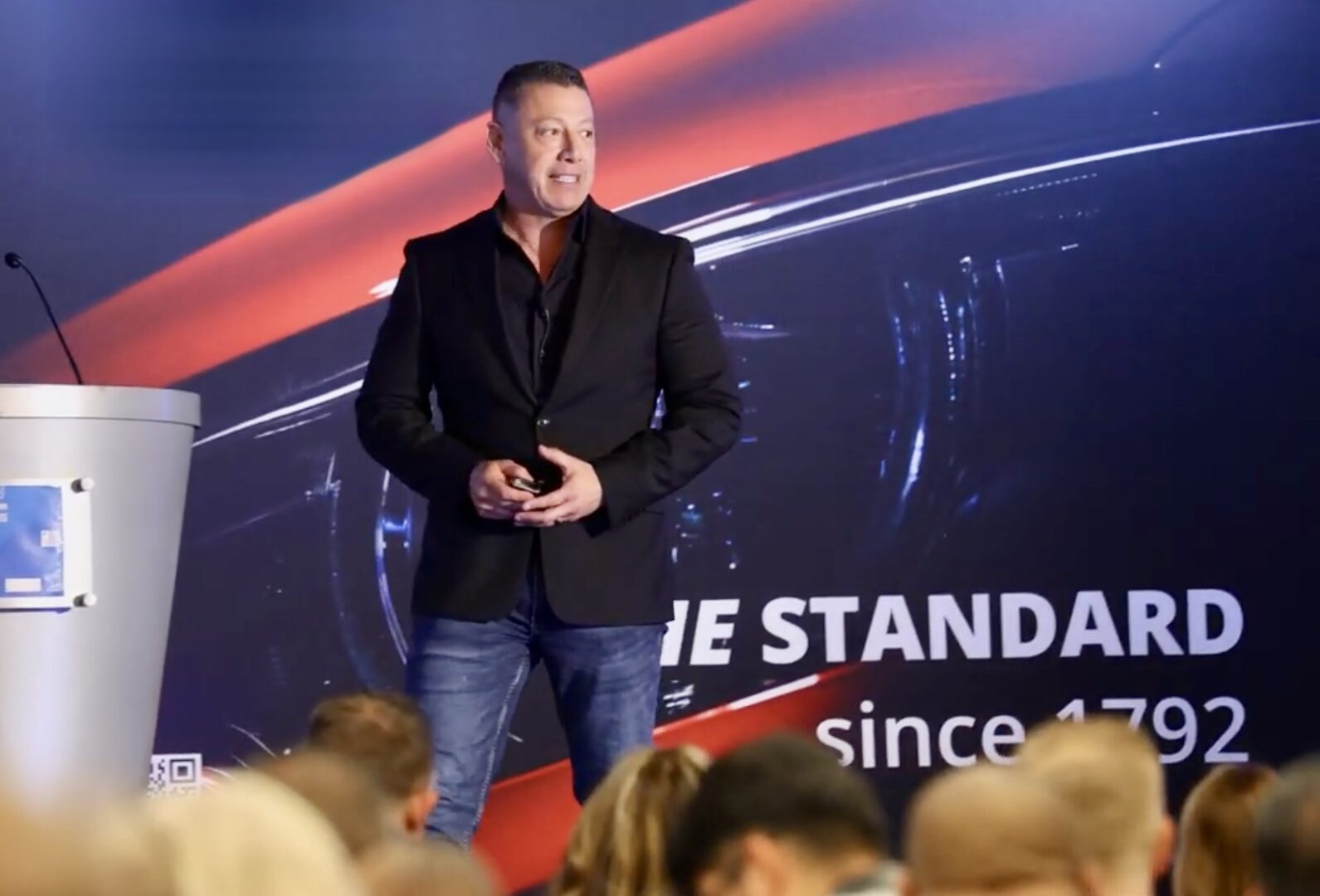 Man in a blazer speaking on stage with an audience.